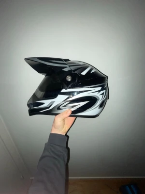 Svartvit crosshjälm med heltäckande visir  - Säljer en svart och vit crosshjälm med coola grafiska mönster och två stycken heltäckande visir framtill. Hjälmen har en skärm upptill och ventilationsöppning vid hakan. Passar perfekt för motocross eller offroad-åkning.Storlek 57-58cm