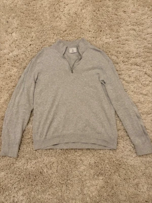 Ljusgrå halvzip tröja från Bläck - Snygg ljusgrå tröja från Bläck med halvzip. Tillverkad i mjukt material med ribbade muddar vid ärmslut och nederkant. Perfekt för en clean och stilren look. Liten i storleken.