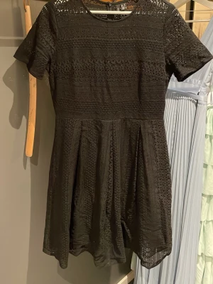 Svart spetsklänning med kort ärm - Säljer en svart kortklänning i spets med rund halsringning och korta ärmar. Klänningen har ett vackert hålmönster och är lätt utställd nedtill för en feminin siluett. Perfekt för dig som gillar detaljer och stilren design.