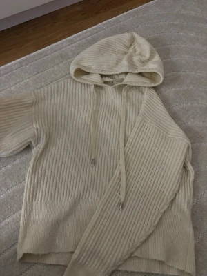 Ribbstickad hoodie från Freequent - Mysig ribbstickad hoodie i krämvit färg från Freequent. Tröjan har huva med dragsko och långa ärmar. Tillverkad i ett mjukt stickat material som känns skönt mot huden. Perfekt för dig som gillar en avslappnad och stilren look.