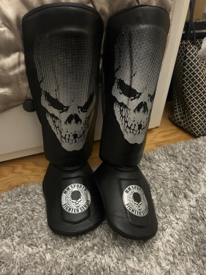Svarta benskydd MM Combat Gear - Säljer ett par svarta benskydd från MM Combat Gear med coolt dödskalletryck framtill och kardborreband för enkel justering. Perfekta för kampsport eller träning där extra skydd behövs. Tillverkade i slitstarkt konstläder.
