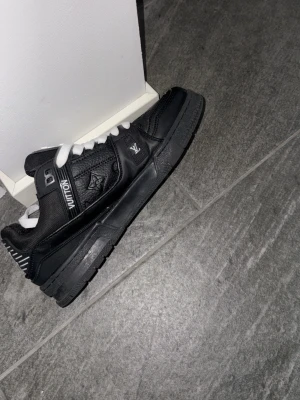 Louis Vuitton svarta sneakers - Svarta Louis Vuitton sneakers i läder med vita detaljer och vita skosnören. Skon har präglade LV-loggor och stjärndetaljer på sidan samt en robust sula. Klassisk low-top modell med exklusiv känsla och tydliga LV-märken på både plös och sidor.