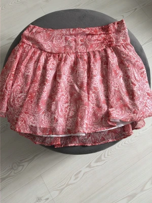 Rosa kortkjol med paisleymönster - Söt kortkjol i rosa/corall och vitt med ett detaljerat paisleymönster. Kjolen har en luftig och volangig form med bred linning och lätt utställd siluett. Perfekt för dig som gillar färg och mönster i garderoben.