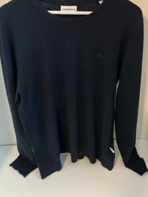 Svart stickad tröja från Lindbergh - Klassisk svart stickad tröja från Lindbergh med rund halsringning och långa ärmar. Tröjan har en diskret logga på bröstet och ribbade muddar vid ärmslut och nederkant. Perfekt basplagg för lager på lager och enkel att matcha.