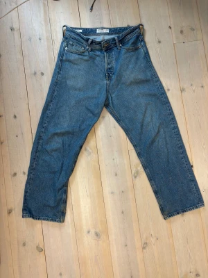 Blå raka jeans från Jack&Jones - Säljer ett par klassiska blå jeans från Jack&Jones med lös passform. Jeansen har fem fickor, bälteshällor och knappgylf. Ett mindre hål vid insidan på ena benet men inget som märks av vid användning. Kan tänka mig gå ner i pris vid snabb affär och tveka inte på att höra av dig vid funderingar!