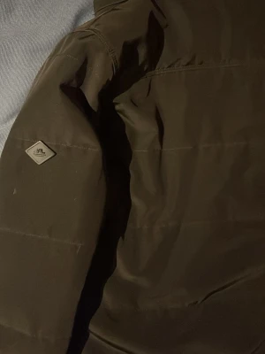 Mörk dunjacka från J.Lindeberg - Snygg mörk dunjacka från J.Lindeberg med diskret logga på ärmen. Jackan är tillverkad i 100% polyester med foder av polyamid och polyesterfyllning. Perfekt för kalla vinterdagar och har en stilren, enkel design.