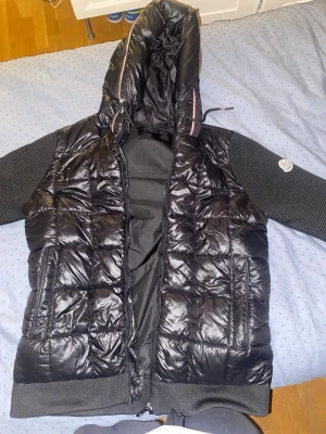 Svart pufferjacka från Moncler - Säljer en svart pufferjacka från Moncler med glansig quiltad front och rygg, stickade svarta ärmar och huva med dragsko. PRIS KAN DISKUTERAS VID SNABB AFFÄR!