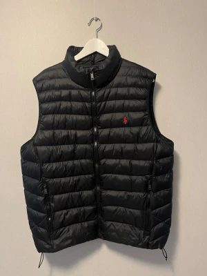 Svart dunväst från Ralph Lauren - Snygg svart dunväst från Ralph Lauren med klassisk röd logga på bröstet. Västen har horisontella sömmar, hög krage och dragkedja framtill samt två fickor med dragkedja. Västen har används väldigt lite och är inprincip i nyskick 