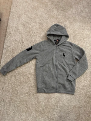 Grå hoodie från Polo Ralph Lauren - Snygg grå hoodie med dragkedja från Polo Ralph Lauren. Klassisk huva med dragsko, fickor framtill och svart broderad logga på bröstet. Siffran 3 i svart på ena ärmen ger en sportig touch. Perfekt för en avslappnad stil. Den är storlek M men passar även någon som bör S