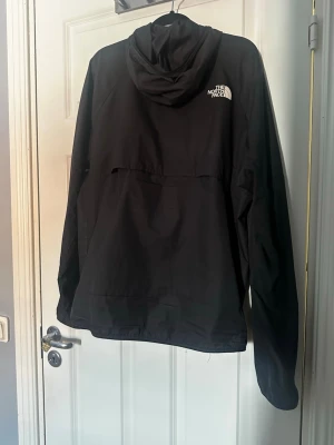 Svart vindjacka från The North Face - Snygg svart vindjacka från The North Face med huva och dragkedja framtill. Jackan har två sidofickor med dragkedja och logga på bröstet och ryggen. Tillverkad i lätt och vindtätt material, perfekt för blåsiga dagar.