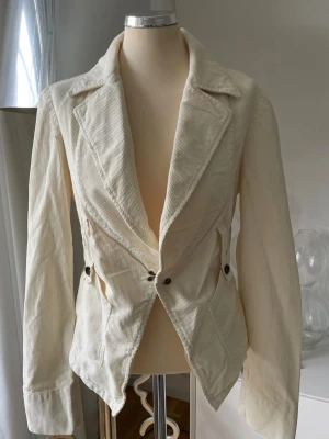 kavaj i manchester från Fornarina - Stilren vit kavaj i manchestertyg med markerad midja och breda slag. Två knappar framtill och dekorativa fickor med knappdetaljer. Långa ärmar med knappslå och sprund bak. Perfekt för dig som gillar klassisk men trendig stil.