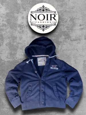 Mörkblå hoodie från Hollister - Snygg mörkblå hoodie från Hollister med knappar framtill och två fickor. Hoodien har en stor huva med vita snören och logotyp på bröstet. Perfekt för dig som gillar en avslappnad och cool stil. 