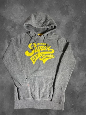 Grå Superdry hoodie med gul text - Snygg grå hoodie från Superdry med stor gul text och logga på bröstet. Klassisk huva med dragsko och magficka framtill. Tillverkad i mjukt sweatshirtmaterial, perfekt för en avslappnad stil. Ribbade muddar vid ärmslut och nederkant.