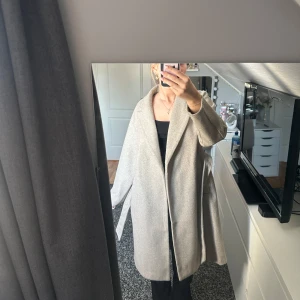 Beige kappa med knytskärp - Säljer en stilren beige kappa med bred krage och knytskärp i midjan. Kappan har en avslappnad passform, långa ärmar och är perfekt för att skapa en snygg look.