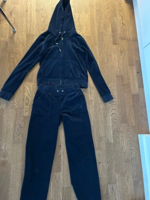 Mörkblå velourset från Juicy Couture - Säljer ett mörkblått velourset från Juicy Couture med luvtröja och byxor. Tröjan har dragkedja, fickor och huva med snörning. Byxorna har bred resår i midjan, fickor fram och bak samt raka ben. Perfekt för chill dagar hemma.