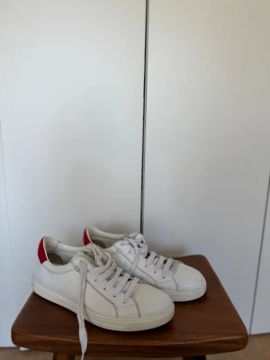  Dsquared2  - Snygga vita sneakers från Dsquared2 med klassisk låg profil och röd detalj på hälen där märkesnamnet syns. Skorna har vita snören och är tillverkade i slätt läder för en clean look. Perfekta för dig som gillar stilrena och trendiga sneakers.
