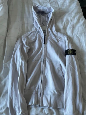 Vit hoodie från Stone Island - Säljer en vit zip hoodie från Stone Island eftersom den tyvär har blivit förliten. Den är köpt på Nk i Stockholm för ett par år sedan och därför självklart äkta! Pris går att diskuteras och är alltid öppen för bytes förslag!