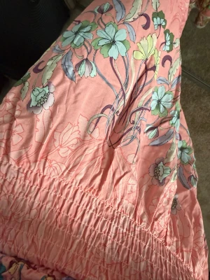 Lång blommig kjol i rosa toner - Säljer en lång kjol i ljusrosa med blommigt mönster i grönt, blått och gult. Kjolen har bred resår i midjan och volangdetaljer nertill. Perfekt för dig som gillar bohemisk och färgglad stil.