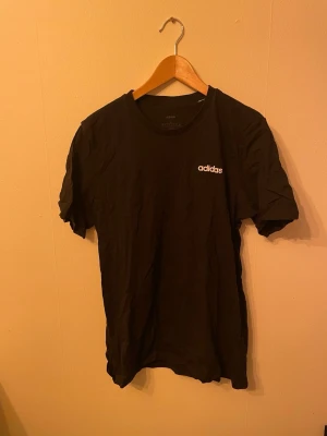 Svart Adidas t-shirt med logga - En svart t-shirt från Adidas med vit logga på bröstet. Klassisk rund hals och korta ärmar. Perfekt för dig som gillar sportiga och stilrena plagg. Materialet är mjukt och känns bekvämt mot huden.