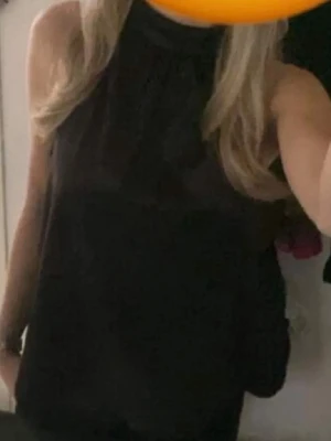 Svart ärmlös halterneck-top - Säljer en svart, ärmlös halterneck-top i glansigt material. Toppen har hög krage och en lös passform som ger en snygg siluett. 