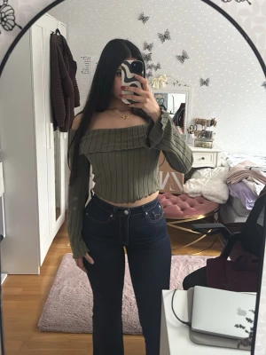Olivgrön ribbad offshoulder topp - Trendig olivgrön croppad topp med ribbad struktur och offshoulder-design. Långa ärmar och bred krage som ger en snygg och modern look. Perfekt för dig som gillar att sticka ut med stilrena detaljer.