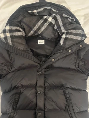 Svart pufferjacka från Burberry - Snygg svart pufferjacka från Burberry med klassiskt rutigt foder i huvan. Jackan har dragkedja och knappar framtill samt två fickor med dragkedja. Perfekt för kalla vinterdagar och riktigt stilren design.