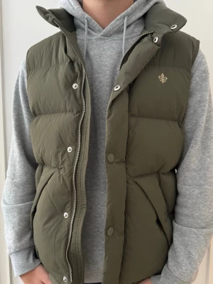 Olivgrön dunväst från Morris - Snygg olivgrön dunväst från Morris med hög krage och diskret logga på bröstet. Västen har dragkedja och tryckknappar framtill samt två stora fickor med lock. Perfekt att slänga över en hoodie för en chill look.