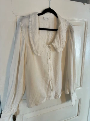 Vit blus med volangkrage från Vila - Supersöt vit blus från Vila med stor volangkrage och knappar framtill. Blusen har långa ärmar och är gjord i ett lätt, krinklat tyg som ger en avslappnad men ändå chic känsla. Perfekt att styla med jeans eller kjol för en trendig look. Passar M/L