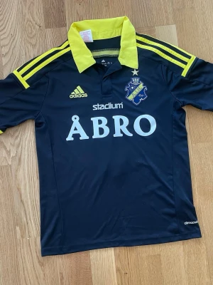 AIK matchtröja, 13-14Y med siffra 5 på ryggen - AIK Fotbollströja från Adidas i svart med gula detaljer. Storlek 164 cm (ca 13-14 år).  Tröjan har AIK-emblemet på bröstet tillsammans med Adidas-logotypen och sponsorloggorna "Stadium" och "ÅBRO". På ryggen finns siffran "5" och texten "SVEA EKONOMI". Kragen är gul och ärmarna har gula ränder. Tröjan är designad med Climacool-teknologi.(Tröjan är i storlek 164 cm och för 13-14 år. Inte XXS, som angetts, jag vet bara inte hur man ändrar till ungdomsstorlek när man lägger in annons). 