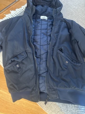 Mörkblå bomberjacka Stone Island - Snygg mörkblå bomberjacka från Stone Island Junior med quiltat foder och mjuk teddykrage. Jackan har två snedställda fickor med knappstängning och breda muddar vid ärmslut och nederkant. Perfekt för dig som vill ha en klassisk och stilren look.