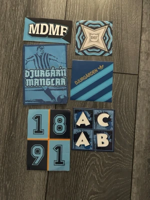 Djurgården klistermärken blå - Säljer ett set med klistermärken med Djurgården-tema i olika blå nyanser och svart. Motiven inkluderar text, siffror och grafiska element, bland annat 'MDMF', 'DIF', 'Djurgården', och '1891'. Perfekt för att dekorera personliga saker.