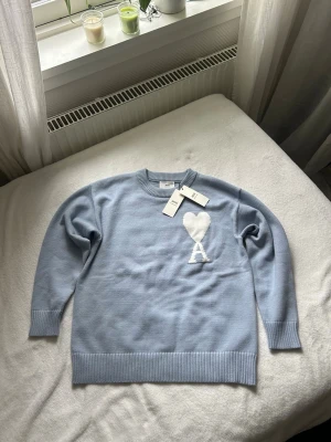 Ljusblå stickad tröja Ami Paris - Supersnygg ljusblå stickad tröja från Ami Paris med deras ikoniska vita hjärta och A broderat på bröstet. Tröjan har rund hals, ribbade muddar och är gjord i mjukt material. Perfekt för dig som gillar stilrena och trendiga plagg.