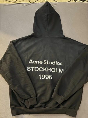 Svart hoodie från Acne Studios - Snygg svart hoodie från Acne Studios med stor huva och tryck på ryggen där det står 'Acne Studios STOCKHOLM 1996'. Klassisk passform med ribbade muddar vid ärmslut och nederkant. Perfekt för dig som gillar stilrena och trendiga plagg.