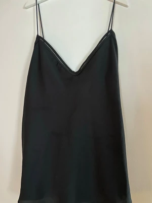 Svart linnetopp med tunna axelband - Stilren svart linnetopp med tunna axelband och v-ringad hals. Toppen har en enkel och clean design med dekorativ söm längs kanten. Perfekt att matcha med jeans eller kjol för en trendig look.