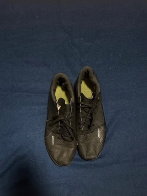 Svarta Adidas fotbollsskor med snörning - Säljer ett par svarta Adidas fotbollsskor med låg profil och snörning. Skorna har en modern design med diskreta ränder och textur på ovandelen. Yttersulan är platt och passar perfekt för spel på inomhusunderlag. Märket syns tydligt på sidan.