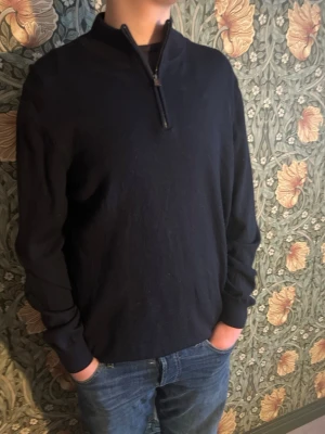 Mörkblå halvzip-tröja 100% ull från BOSS - Står XL men är mer som en Large. Snygg mörkblå stickad tröja från BOSS med halv dragkedja och hög krage. Tröjan har ribbade muddar vid ärmslut och nederkant, perfekt för en clean och stilren look. Materialet känns mjukt och bekvämt, och den passar lika bra till jeans som till chinos.