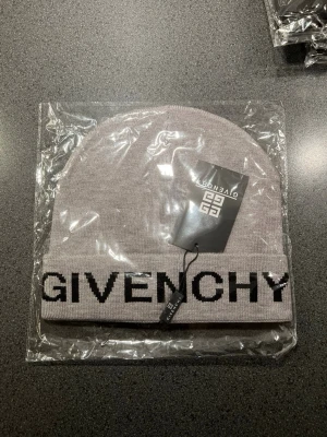 Grå mössa från Givenchy - Säljer en snygg grå mössa från Givenchy med uppvikt kant och stor svart logotyptext framtill. Mössan är stickad och har en clean, modern look som passar perfekt till streetwear och trendiga outfits.