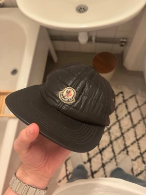 Svart Moncler keps med vaddering - Väldigt väldigt sällsynt keps, inte såld sedan 2019. Visserligen lite sliten men inte många exemplar av denna finns kvar på marknaden.  Säljer en svart keps från Moncler med vadderad, quiltad design och klassisk böjd skärm. Framtill sitter Monclers ikoniska logotyp. Justerbar rem bak med färgdetaljer i rött, gult och grönt. Perfekt för dig som vill ha en unik och snygg accessoar.