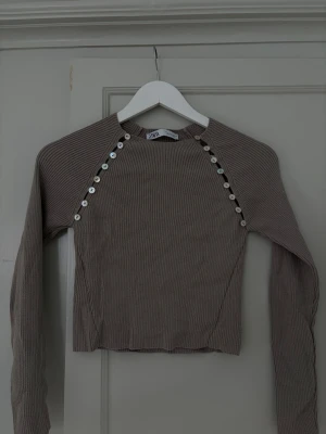 Beige ribbad tröja från Zara - Säljer en beige ribbstickad tröja från Zara med långa ärmar och dekorativa knappar längs axlarna. Tröjan har en kroppsnära passform och rund halsringning. Perfekt för dig som gillar stilrena och trendiga plagg.