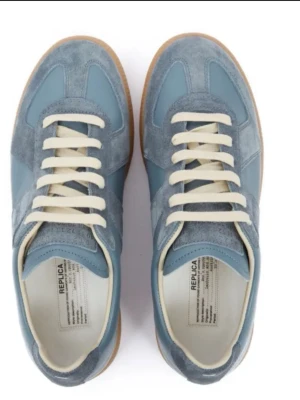  Margiela Replica sneakers - Snygga blå Maison Margiela Replica sneakers med ovandel i mocka och läder, vita snören och klassisk gummisula. Skorna har en stilren low-top design och ikonisk etikett på plösen. Perfekta för dig som gillar minimalistisk och trendig stil.