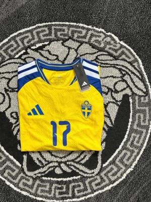 Svensk landslagströja Adidas gul - Säljer en gul svensk landslagströja från Adidas med blå detaljer och nummer 17 på framsidan. Tröjan har korta ärmar, broderad landslagssköld och klassiska Adidas-ränder på axlarna. Perfekt för fotbollsfans som vill visa sitt stöd.