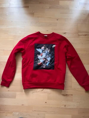 Röd sweatshirt med tryck från Nike - Röd sweatshirt från Nike med ett stort svart tryck framtill som visar sneakers, blommor och duvor. Tröjan har rund halsringning och ribbade muddar vid ärmslut och nederkant. Perfekt för dig som gillar streetwear och snygga detaljer.