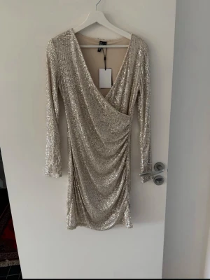 Glittrig omlottklänning  - Säljer en glittrig kort klänning från vero Moda i champagnefärg med omlott och v-ringning. Klänningen har långa ärmar och är täckt av paljetter som ger ett riktigt statement-utseende. Perfekt för dig som vill sticka ut och glänsa.