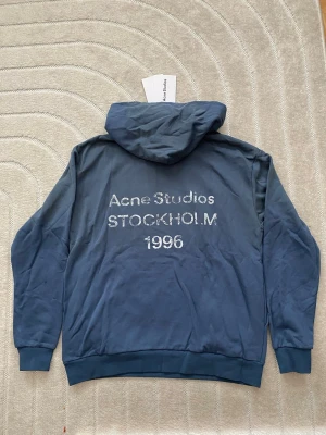Blå hoodie från Acne Studios - Snygg blå hoodie från Acne Studios med tryck på ryggen där det står 'Acne Studios STOCKHOLM 1996'. Hoodien har huva, långa ärmar och ribbade muddar. Tillverkad i mjukt bomullsmaterial och har en clean look utan fickor framtill.