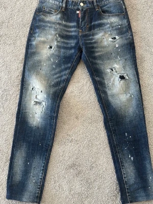 Blå distressed jeans från Dsquared2 - Säljer ett par blå jeans från Dsquared2 med slitna detaljer, hål och färgstänk för en riktigt edgy look. Jeansen har klassisk femficksmodell, knappgylf och läderpatch bak i midjan. Perfekta för dig som gillar streetstyle och vill sticka ut.
