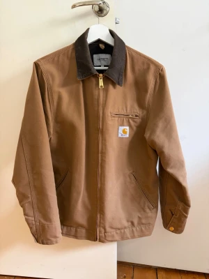 Brun Carhartt workwear jacka - Klassisk brun workwear jacka från Carhartt med mörkbrun krage i manchester. Jackan har dragkedja framtill, tre fickor och Carhartt-logga på bröstet. 