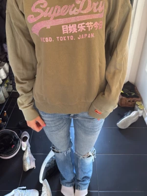 Olivgrön sweatshirt från Superdry - Olivgrön sweatshirt från Superdry med rosa tryck och text på bröstet. Tröjan har rund halsringning, ribbade muddar vid ärmslut och nederkant samt en avslappnad passform. Perfekt för en casual och sportig stil.