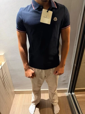 Momcler pike - Snygg marinblå pikétröja från Moncler med klassisk krage och korta ärmar. Tröjan har diskreta röda och vita ränder på krage och ärmslut samt Moncler-logga på bröstet. Perfekt för en stilren och sportig look. Finns svart, mörkblå, vit alla storlekar 