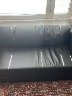 Svart bäddsoffa i konstläder - Säljer en svart bäddsoffa med stilrena raka armstöd och synliga vita sömmar. Soffan är klädd i konstläder och kan enkelt fällas ut till en säng. Perfekt för dig som vill ha en smidig lösning för extra sovplats.