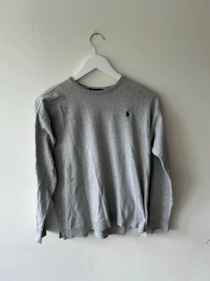 Grå tröja från Polo Ralph Lauren - Klassisk grå långärmad tröja från Polo Ralph Lauren i mjuk pima bomull. Tröjan har rund halsringning och den ikoniska lilla broderade loggan på bröstet. Enkel och stilren design som passar till det mesta.
